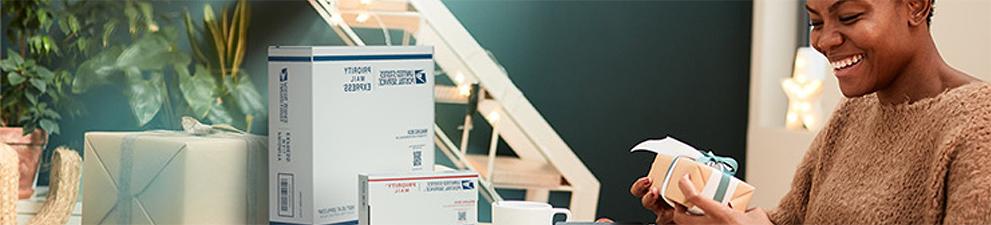 A woman on a laptop preparing to ship Priority Mail boxes and a card with a holiday-themed stamp.